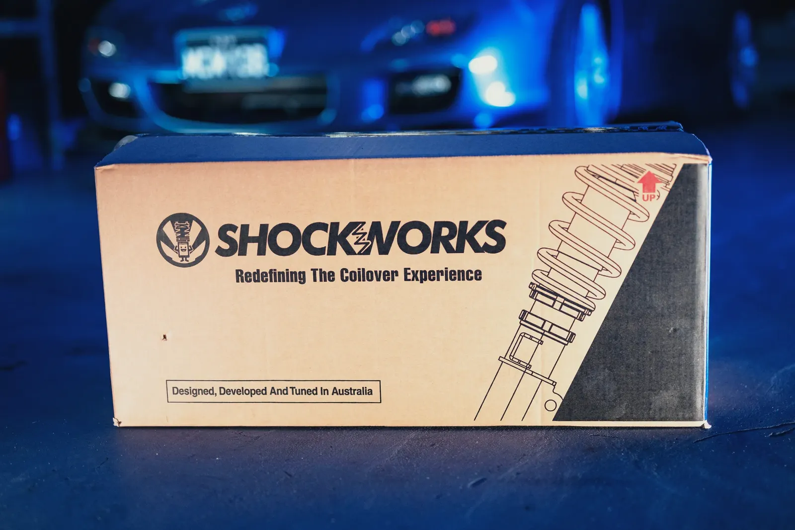 A Shockworks coilover box on the workshop floor, a blue Mazda RX-7 soft in the background