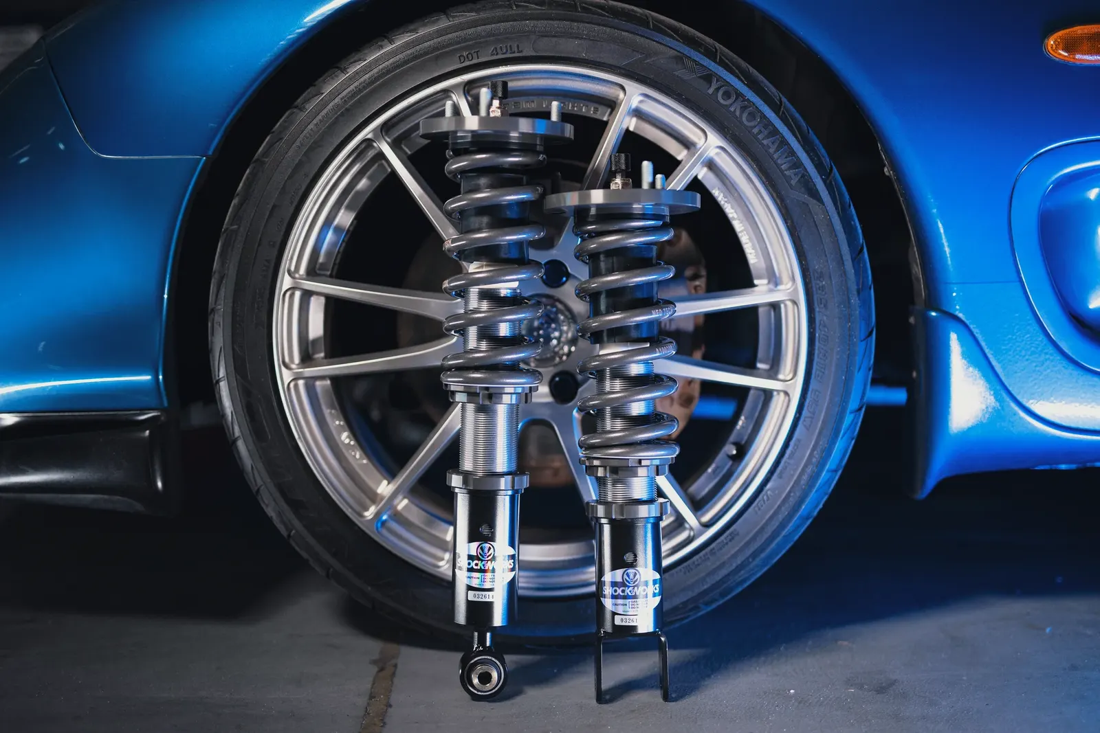 A pair of assembled Shockworks coilovers standing against the alloy wheel of a blue Mazda RX-7