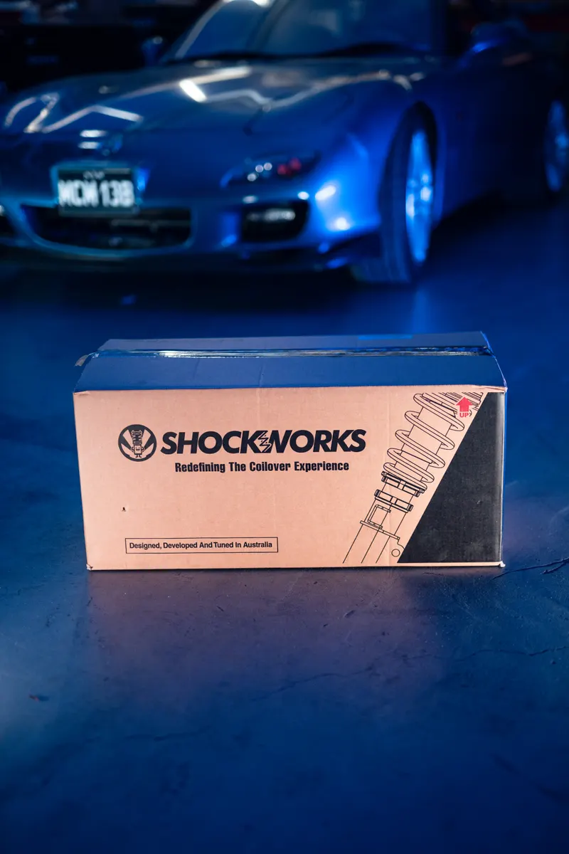 A Shockworks coilover box on the workshop floor, a blue Mazda RX-7 soft in the background