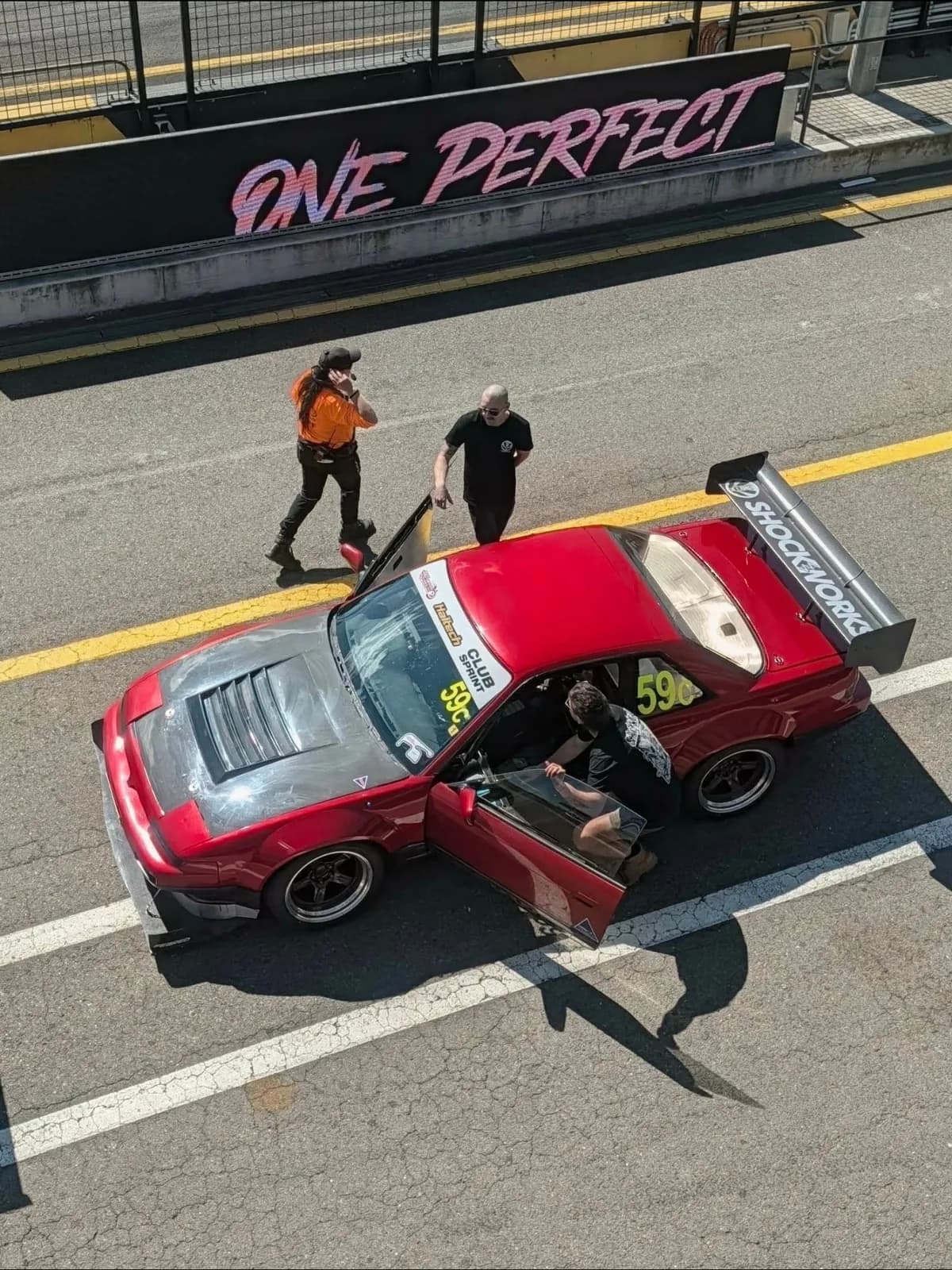 The Shockworks S13 #59c from above in pit lane at Sydney Motorsport Park, door open and the driver climbing in, crew alongside, the One Perfect Lap sign on the wall behind