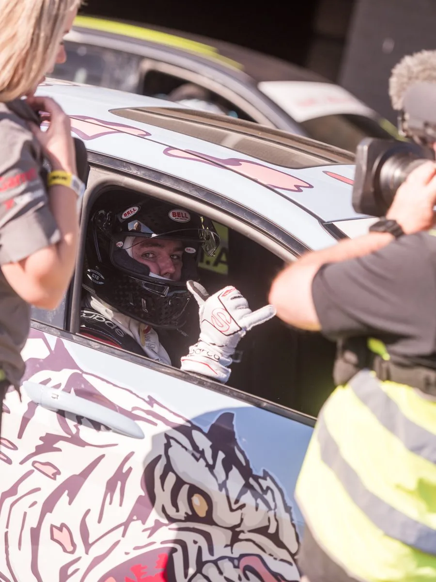Andrew Price helmeted in a race car's driver seat, shaka out the window, with a video camera trained on him