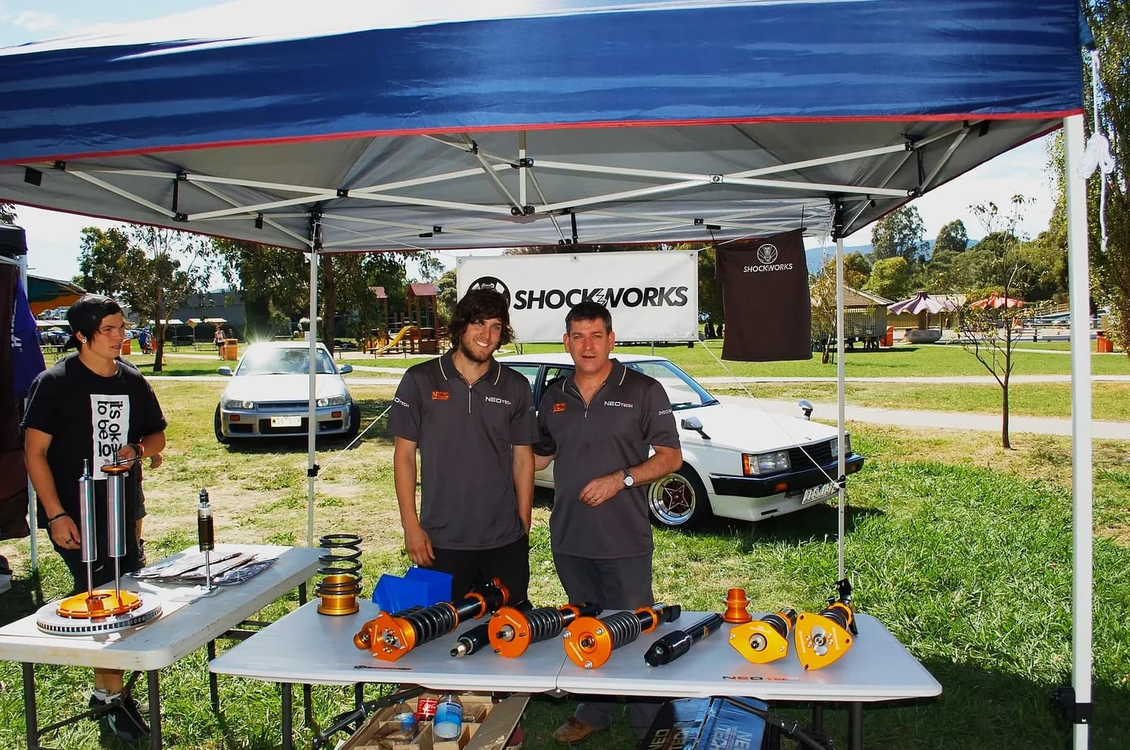 Three men under a blue Shockworks pop-up tent at an outdoor event, coilovers laid out on the tables in front of them