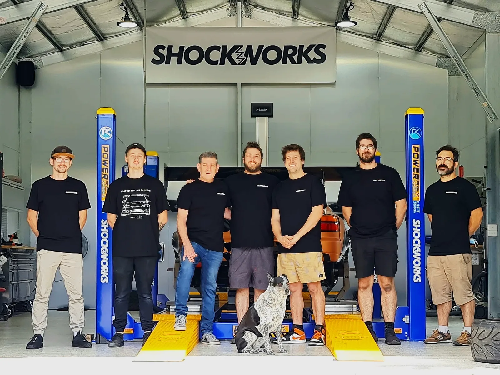 Seven of the crew in matching black Shockworks shirts on the hoist bays under the big Shockworks sign in the workshop