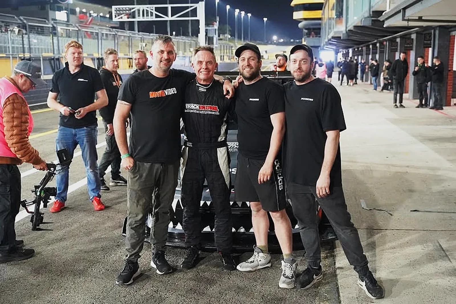 Five men in black Shockworks shirts posing in pit lane at night, one in a race suit, a race car parked behind them