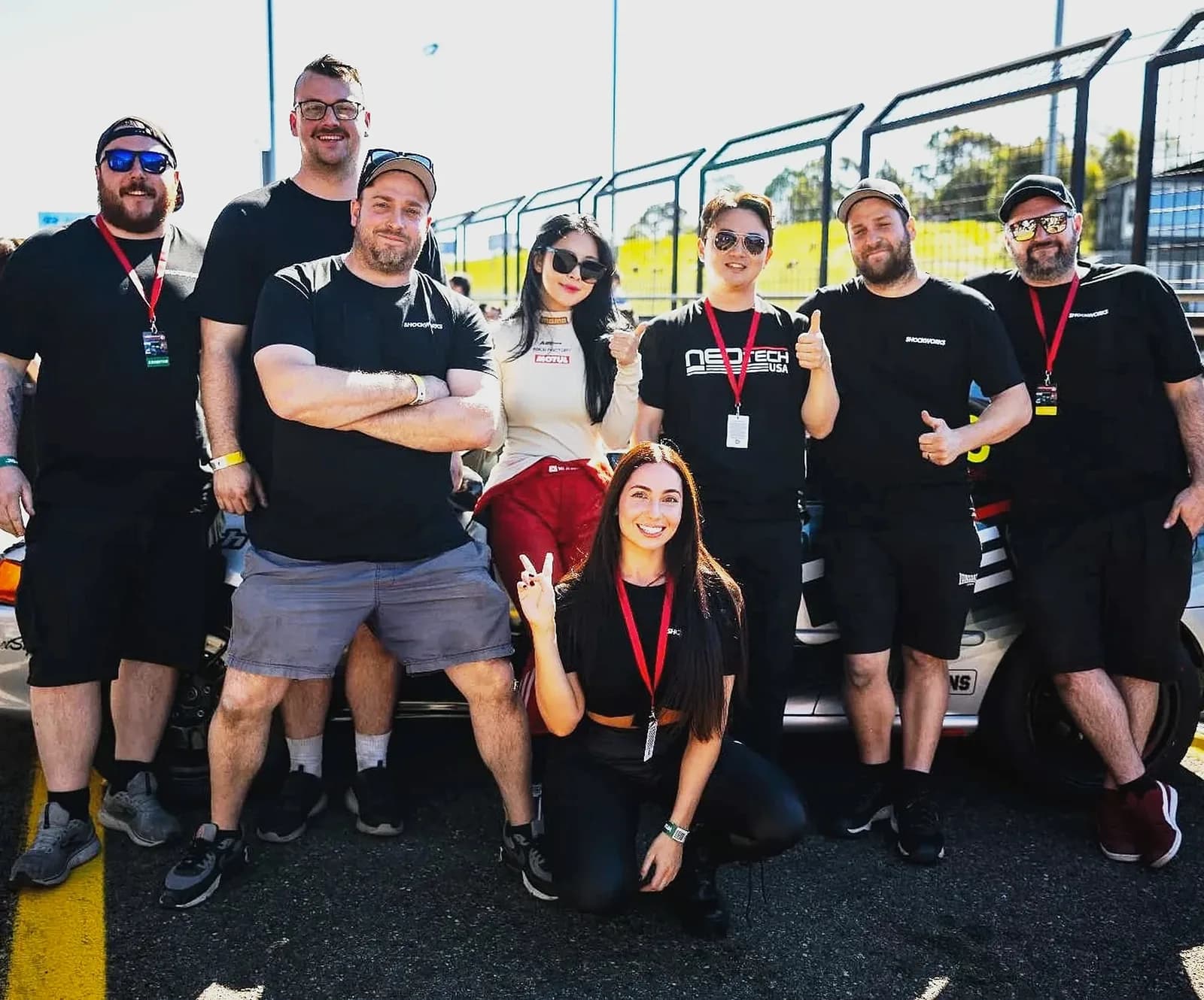 Eight people in black shirts and lanyards posing in pit lane in front of a race car at Sydney Motorsport Park