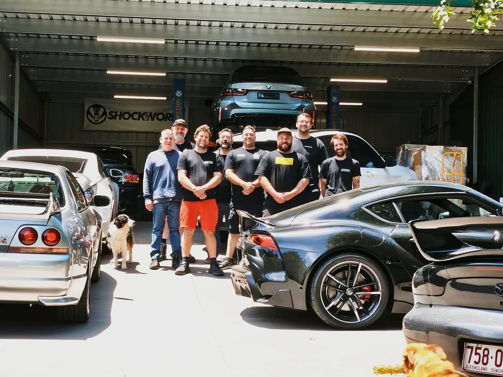 Eight of the crew on the driveway out the front of the Monbulk shed, an R33 and a Supra parked either side of them