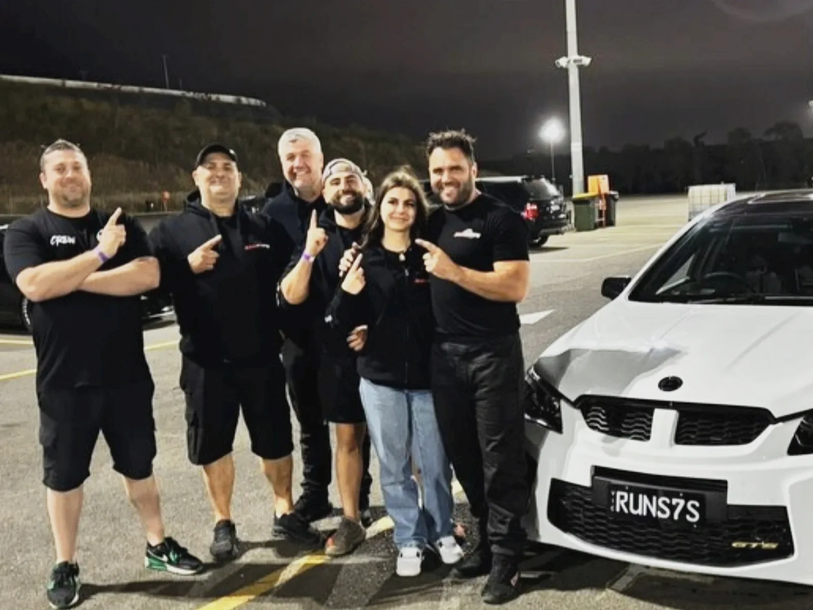 Six of the crew in black shirts posing at night beside a white HSV GTS sedan in a car park