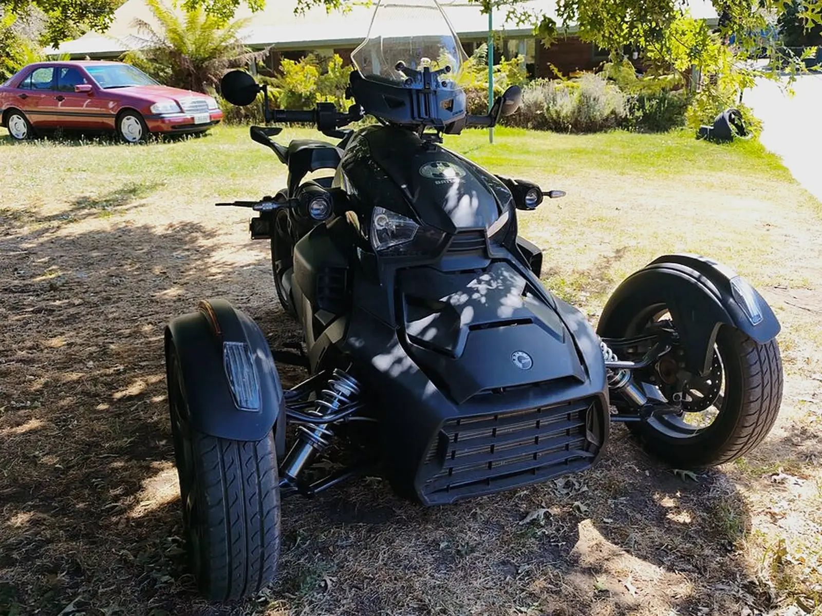 A matte black Can-Am three-wheeler on a lawn, front coilovers visible on both front wheels