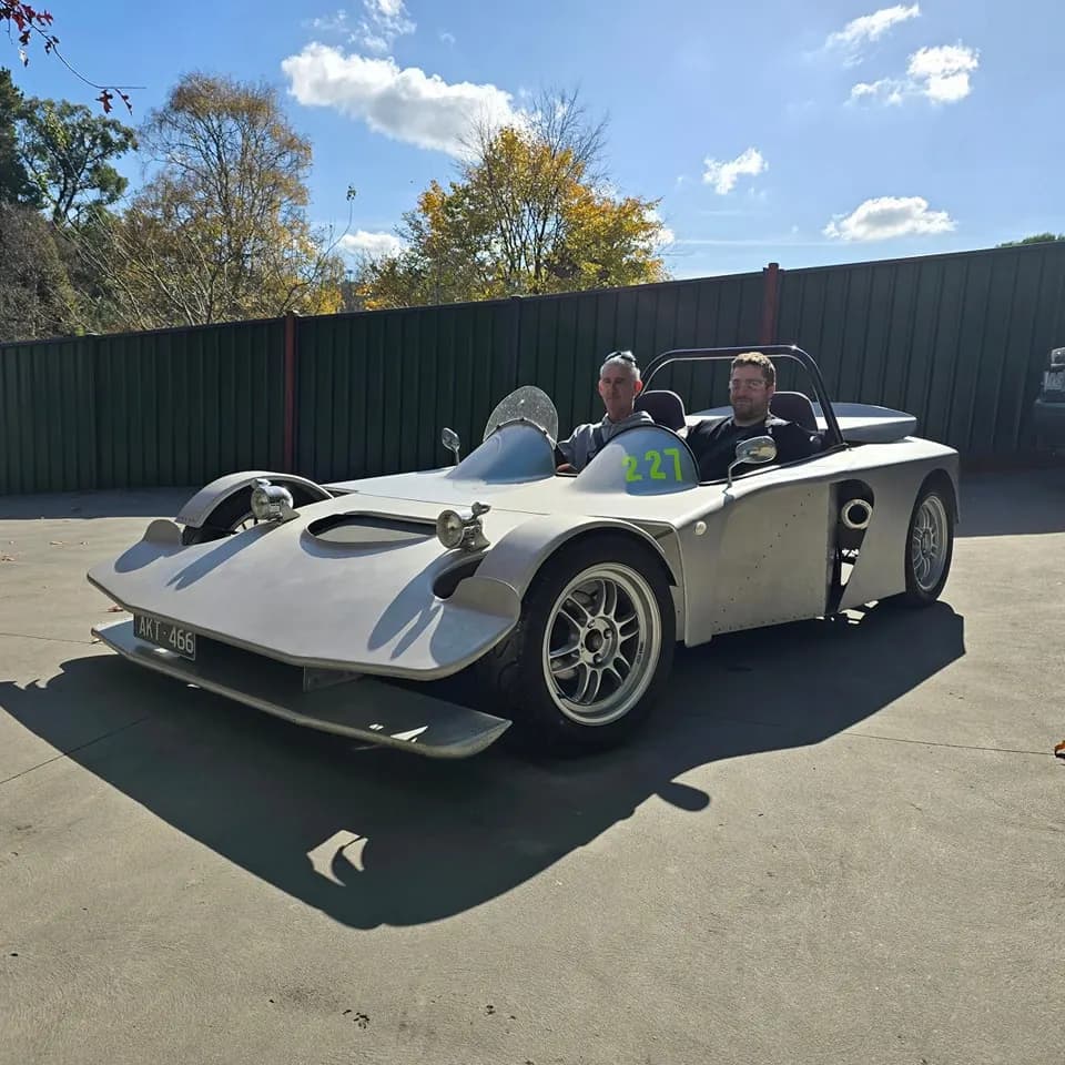 Two men in the bare-aluminium open-top, race number 227, parked in the sun outside the workshop
