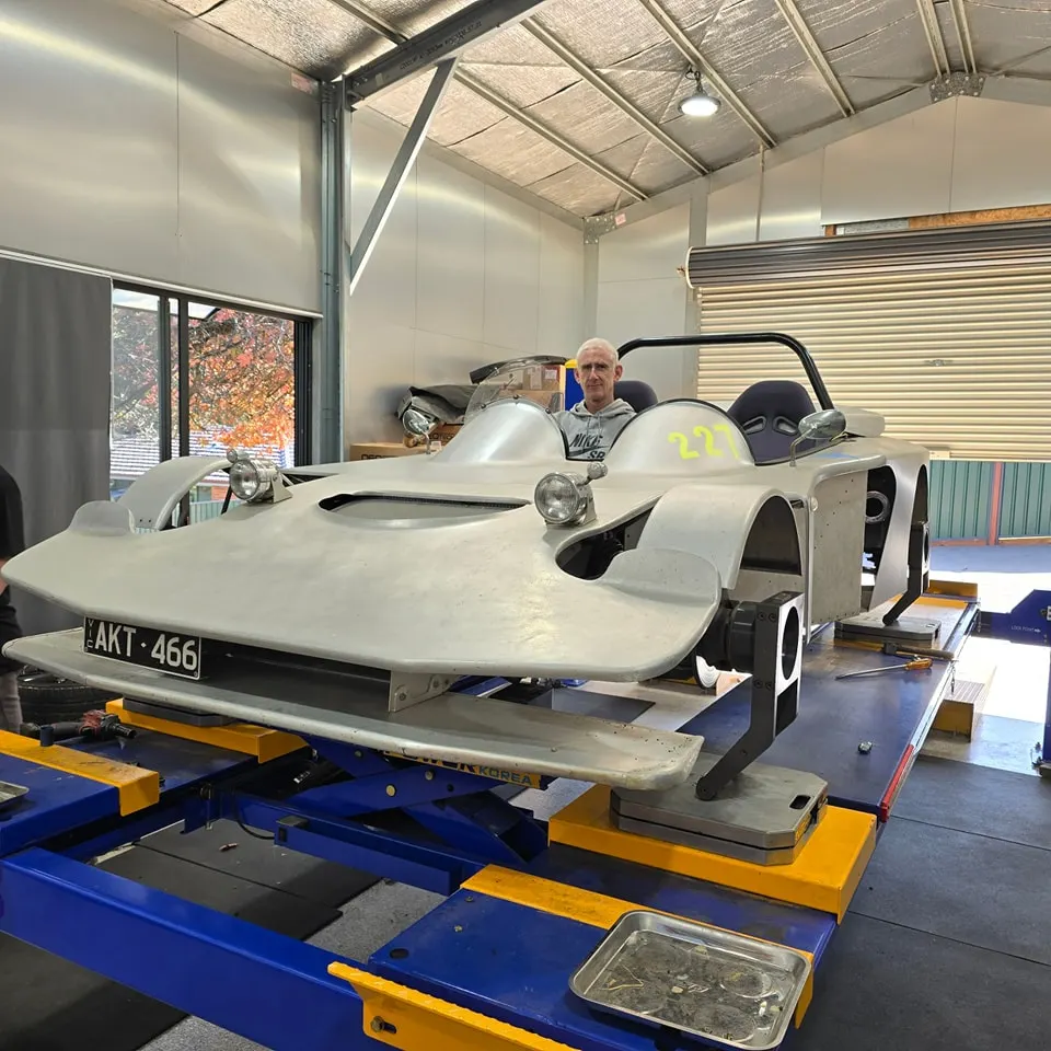 The bare-aluminium open-top front-on on the lift, headlights fitted, a man standing behind the cockpit
