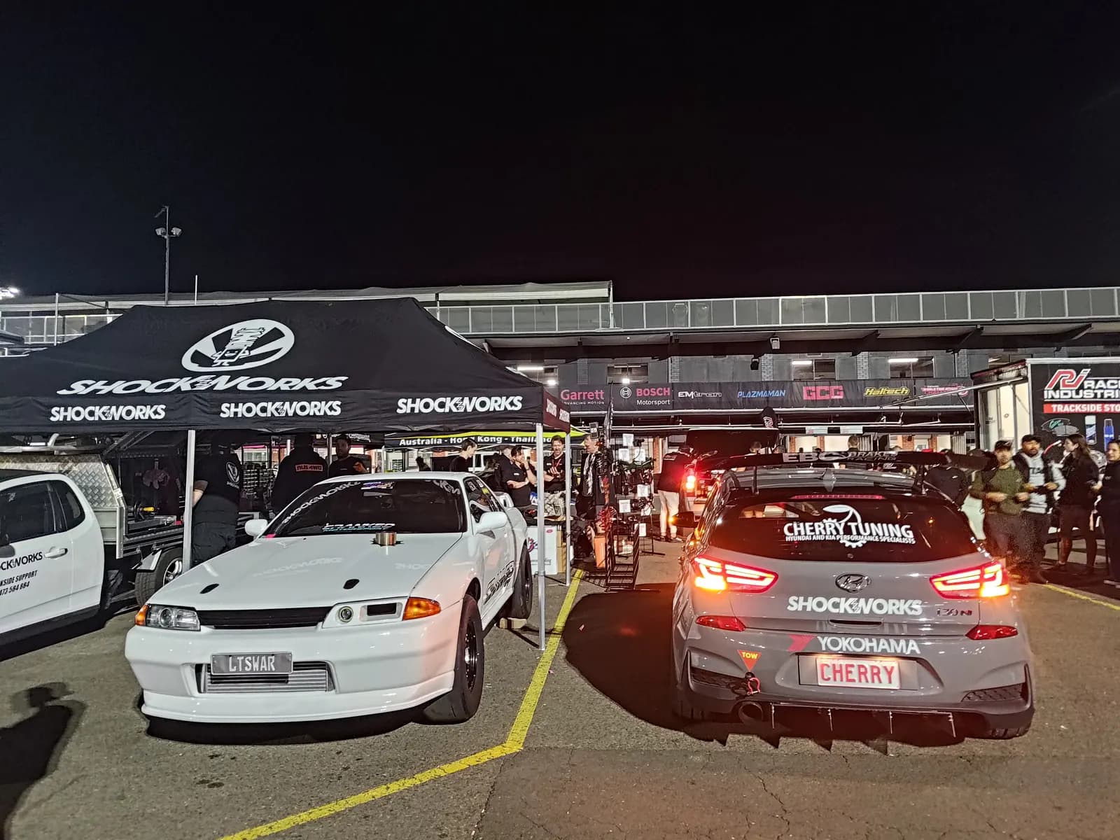 Two Shockworks marquees over customer cars in a pit paddock at night, a white R32 Skyline and an i30N wearing Shockworks decals