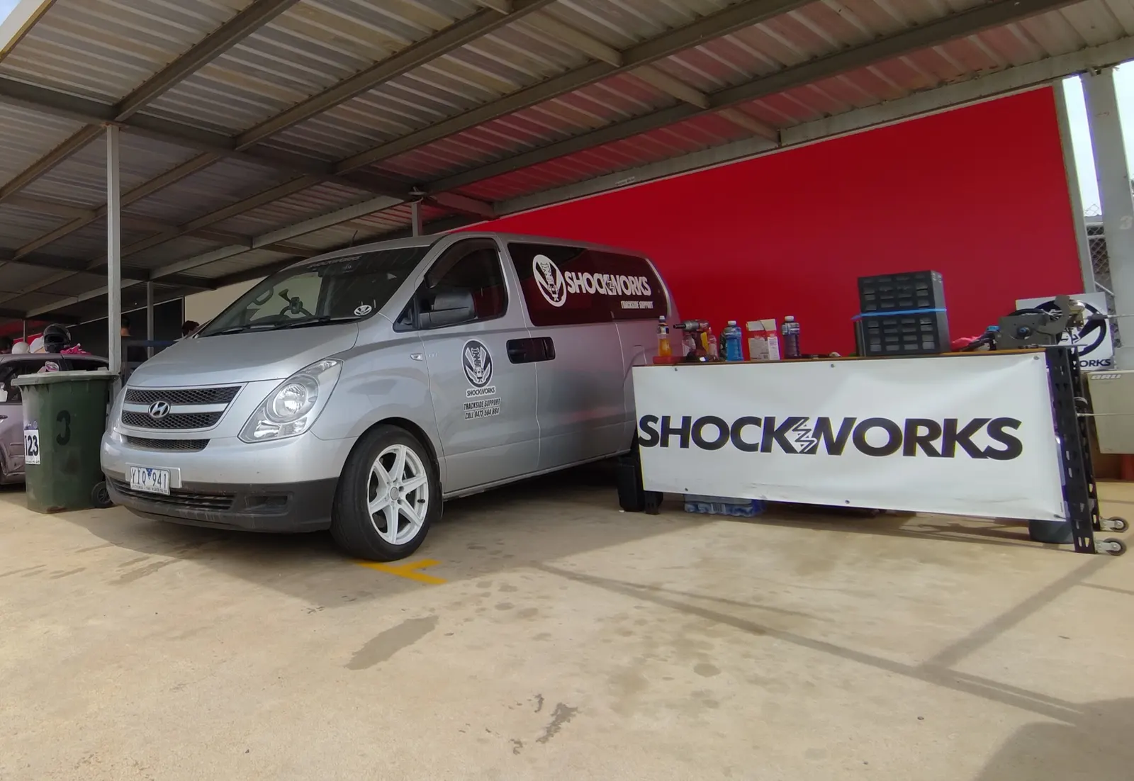 The Shockworks-liveried van parked in a circuit garage bay beside a bench with a Shockworks banner