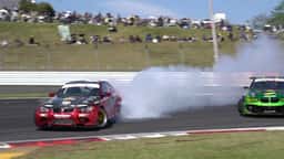 Two drift cars in tandem at a circuit in daylight, the lead car trailing tyre smoke across the track with spectators on the grass bank behind